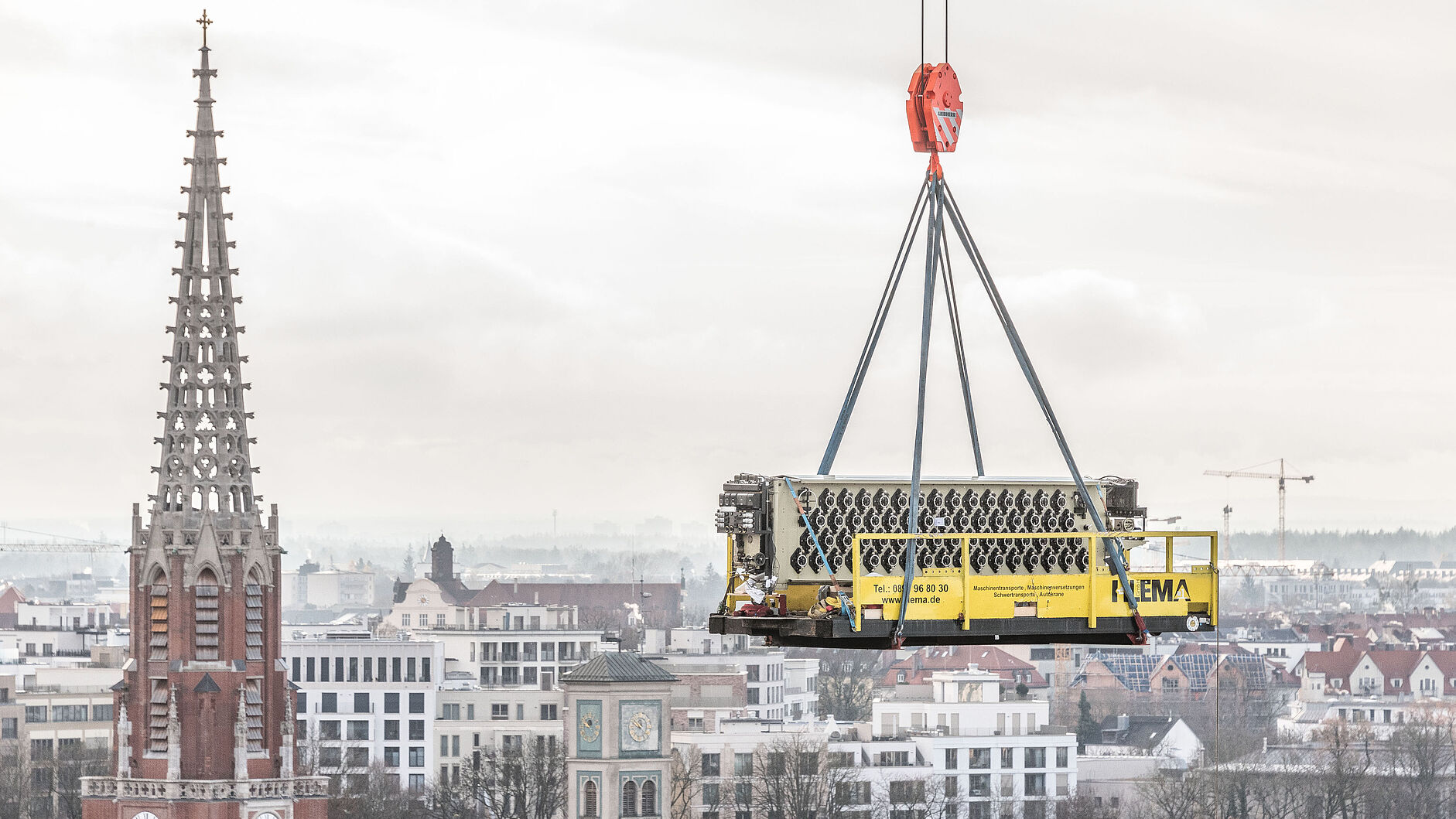 Ein gelber Lastkorb mit großem Maschinenaggregat, aufgehängt an vier Stahlseilen und einem roten Kranhaken, schwebt über einer Stadtlandschaft mit markanter gotischer Kirchturmspitze und Uhrturm im Vordergrund.