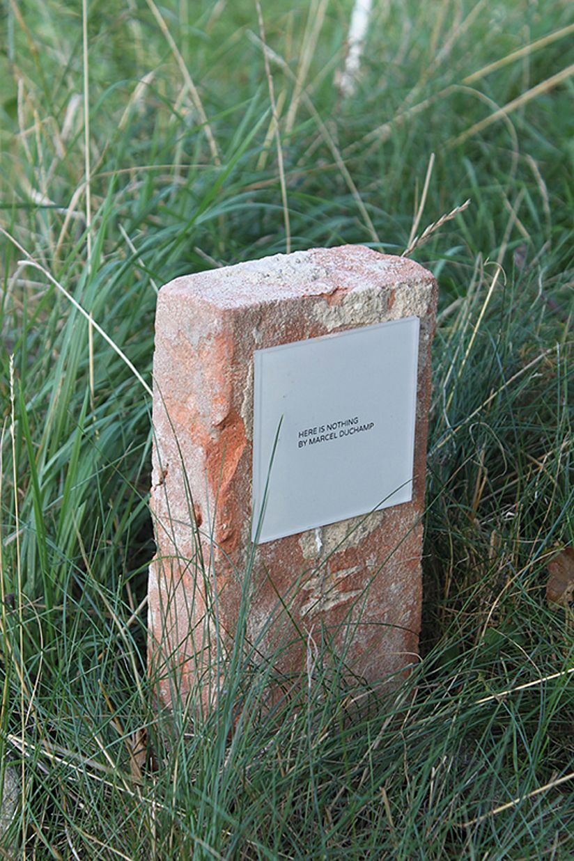 Rechteckiger Stein in einer Wiese. Auf dem Stein befindet sich eine Tafel mit einer Aufschrift in Großbuchstaben "Here is nothing. Marcel Duchamp".