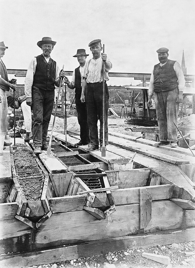 Schwarz-weiß Fotografie. Sechs Bauarbeiter auf einer Holzkonstruktion, einer etwas abseits rechts, die anderen in einer Gruppe und jeweils mit einem Stock in der Hand, alle Blick in die Kamera. Herstellung von Eisenbetonpfählen mit einem Holzgerüst.