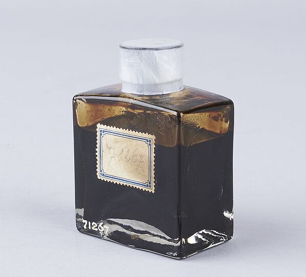 Glasflasche mit dunklem, zähflüssigem Erdöl gefüllt. Die viereckige Flasche hat einen weißen Verschluss und ein Etikett auf der Vorderseite.