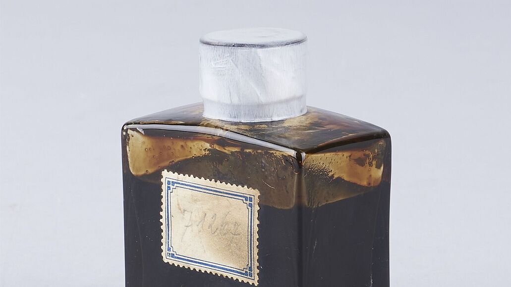 Glasflasche mit dunklem, zähflüssigem Erdöl gefüllt. Die viereckige Flasche hat einen weißen Verschluss und ein Etikett auf der Vorderseite.