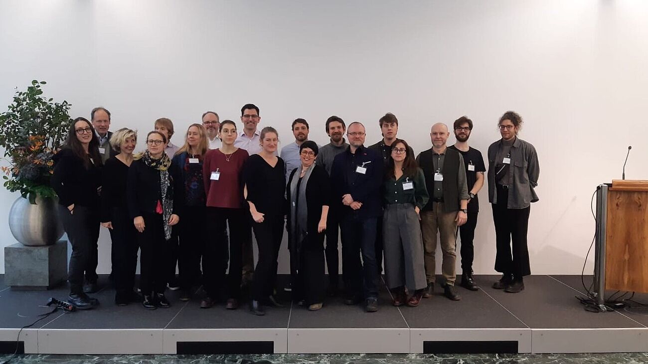 Eine Gruppe von ca. 20 Personen steht leicht erhöht auf einem Podium. Rechts sieht man ein Sprecherpult, links eine Pflanze. 