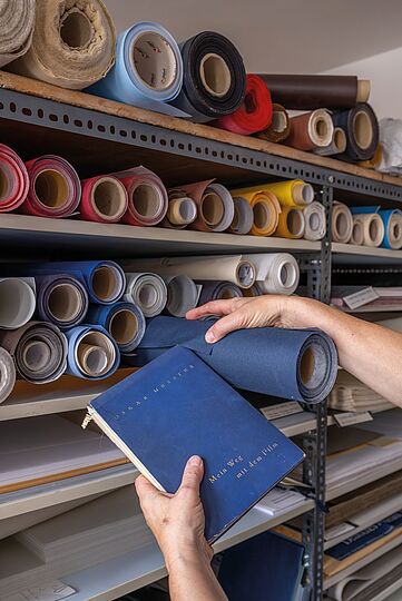 Fotografie. Ein Buch wird vor ein Regal mit verschieden farbigen Rollen gehalten. Eine Hand zieht eine ähnlich Farbe wie die des Einbands des Buches aus dem Regal