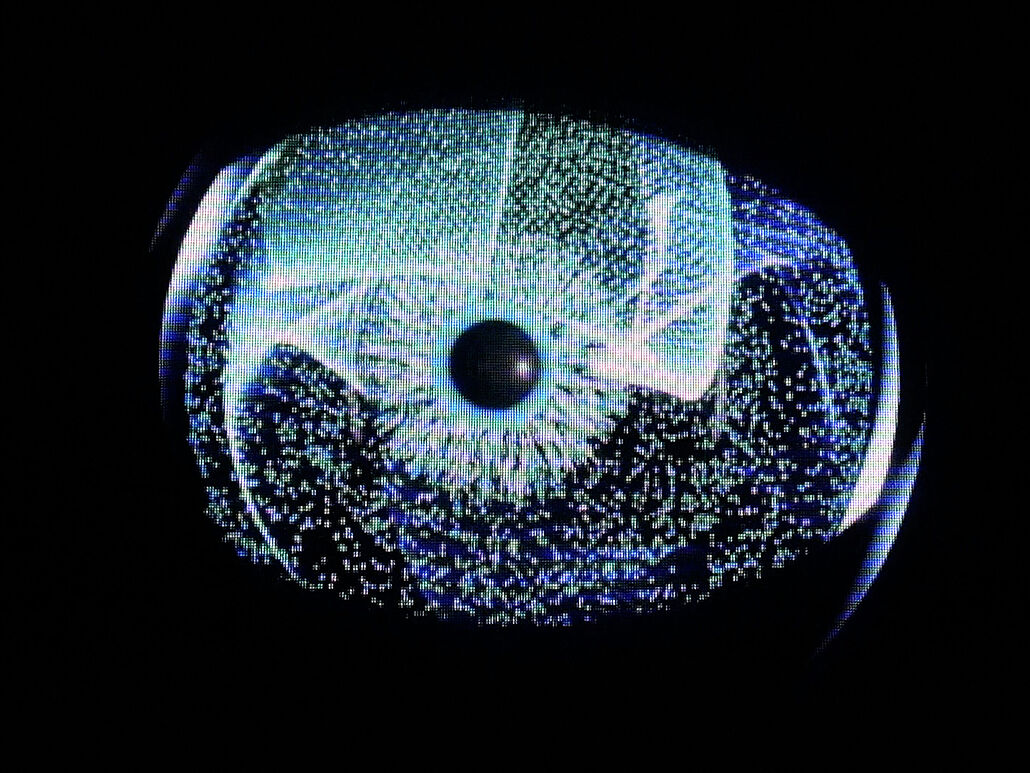 Nahaufnahme einer leuchtenden, digitalen Iris-ähnlichen Grafik: zentrale dunkle Pupille, radial strahlende blaue und grüne Pixelpunkte und Linien auf schwarzem Hintergrund, erinnert an ein Auge oder ein abstraktes Datavisualisierungs-Interface.
