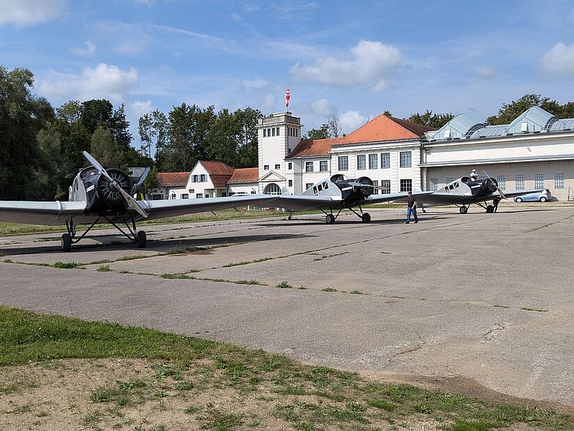 Drei silberne Propellermaschinen stehend auf Betonfläche. Längliches Gebäude mit Türmchen im Hintergrund.