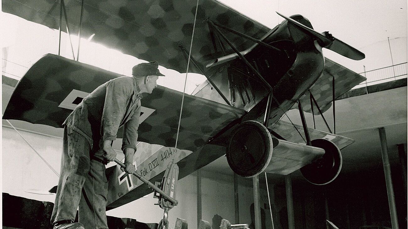 Ein schwarweißes Foto eines historischen Doppeldeckerflugzeugs (Fokker D.VII) hängend in einer Halle, während ein Arbeiter in Arbeitskleidung und Mütze mit einem Hebel an einer Takelvorrichtung steht, um das Flugzeug zu positionieren; sichtbare Details: Propeller, große Speichenräder, Tarnmuster unter dem oberen Flügel und die Kennzeichnung "Fok. D.VII 4404/15" am Rumpf.