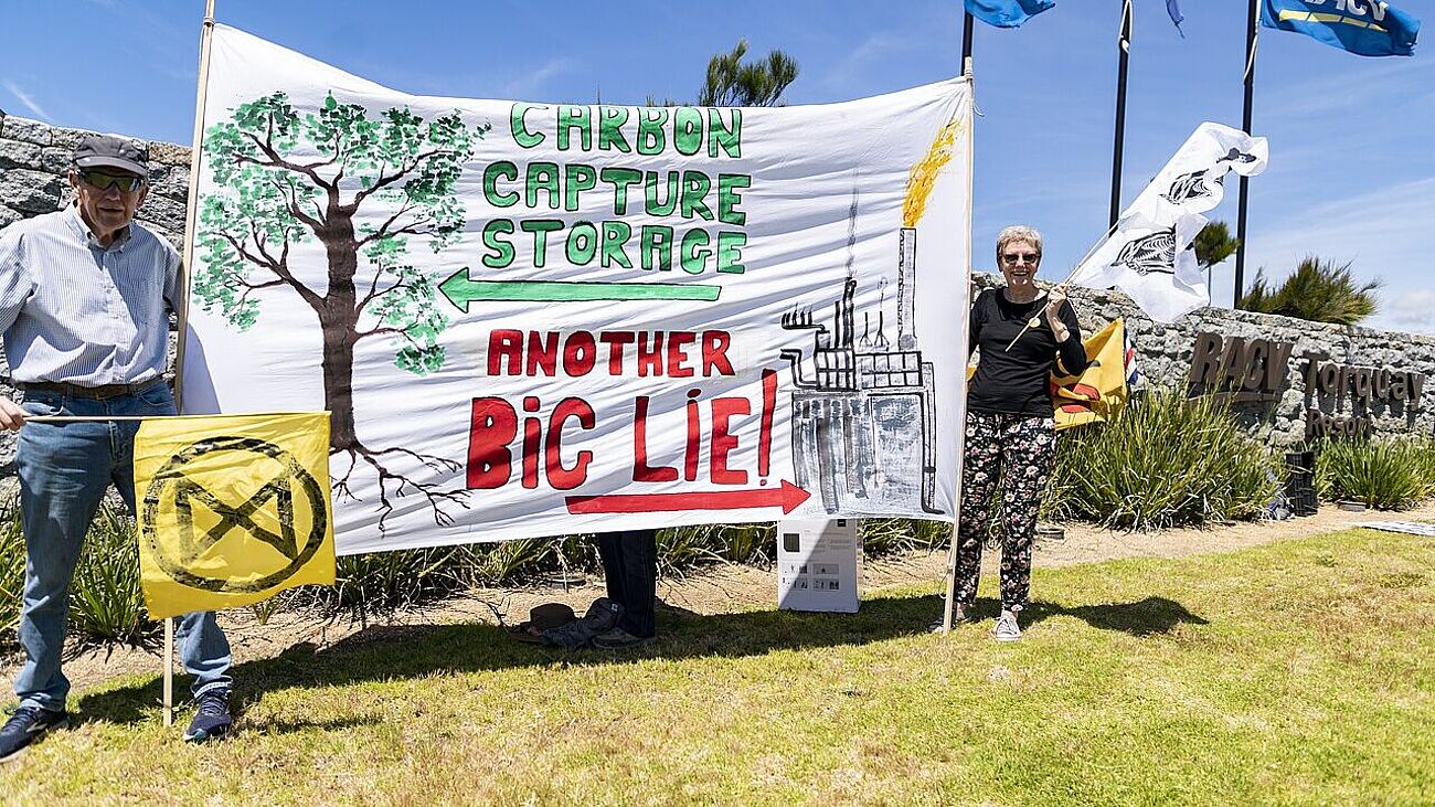 Drei Demonstrierende halten ein großes handgemaltes Transparent mit der Aufschrift „CARBON CAPTURE STORAGE“ und „ANOTHER BIG LIE!“, links ein gemalter Baum, rechts eine Fabrik mit rauchendem Schornstein; gelbe Extinction-Rebellion-Fahne links, weiße Protestfahne rechts, Grasfläche und Steinmauer im Hintergrund unter blauem Himmel.