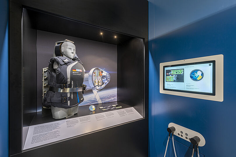 Vitrine in einem Museumsausstellungsraum mit einer Schutzweste auf einem Torso-Display, daneben Modellteile, Infotafeln und ein beleuchtetes Hintergrundbild einer Raumkapsel über der Erde; rechts an der blauen Wand ein eingebautes Touchscreen-Informationsdisplay und darunter ein Bedienfeld mit zwei Hörern.
