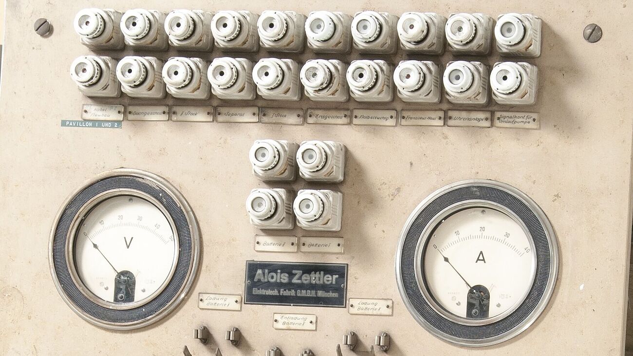 Schalttafel auf Europalette. Sicherungen, Volt- und Amperemeter, verschiedene Hebel, Knöpfe und Bedienelemente in Schwarz. Alles auf heller Holzplatte.
