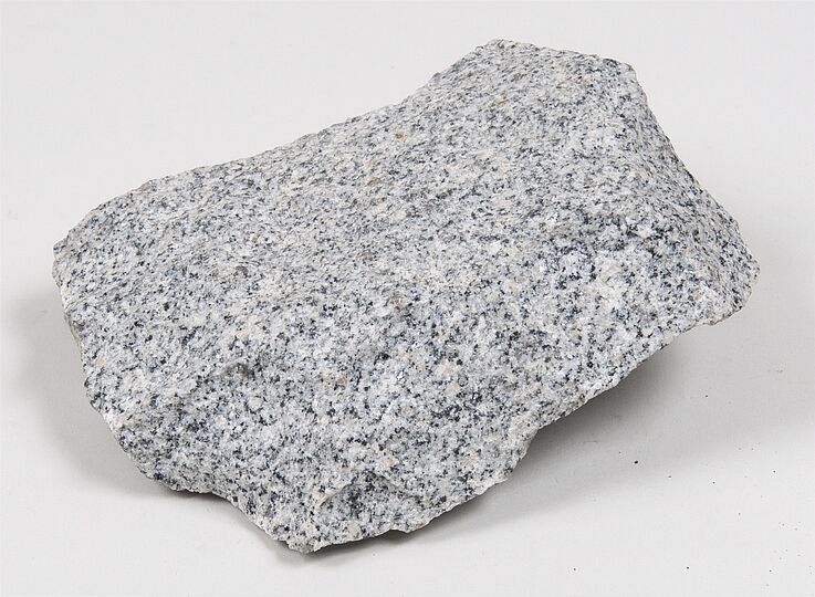 Graues Granitstück mit typischer, grobkörniger Struktur. Das Gestein zeigt ein gesprenkeltes Muster aus hellen und dunklen Mineralien und hat unregelmäßige Bruchkanten.