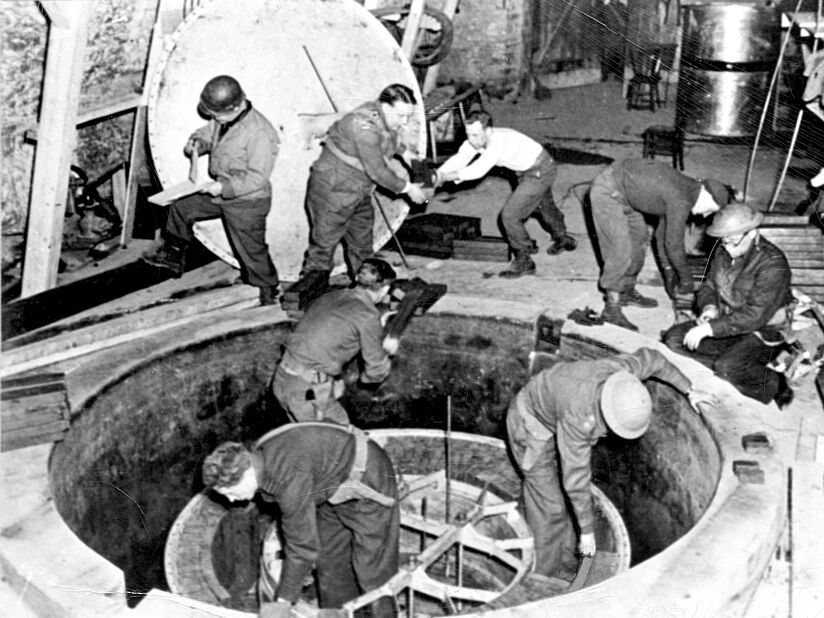 Fotografie. Acht Männer in Uniform, teils mit Helm, verrichten Arbeiten an einem im Boden eingelassen ca. 2 Meter breiten Apparat.