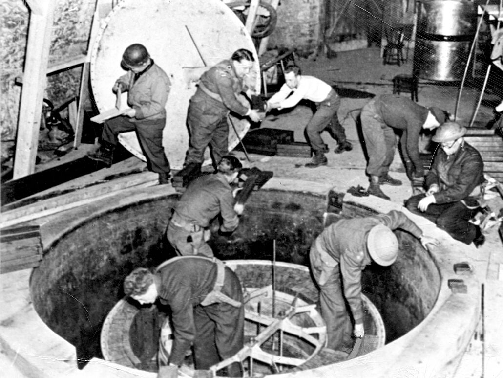 German Uranium Machine 1945 Fotografie. Acht Männer in Uniform, teils mit Helm, verrichten Arbeiten an einem im Boden eingelassen ca. 2 Meter breiten Apparat.