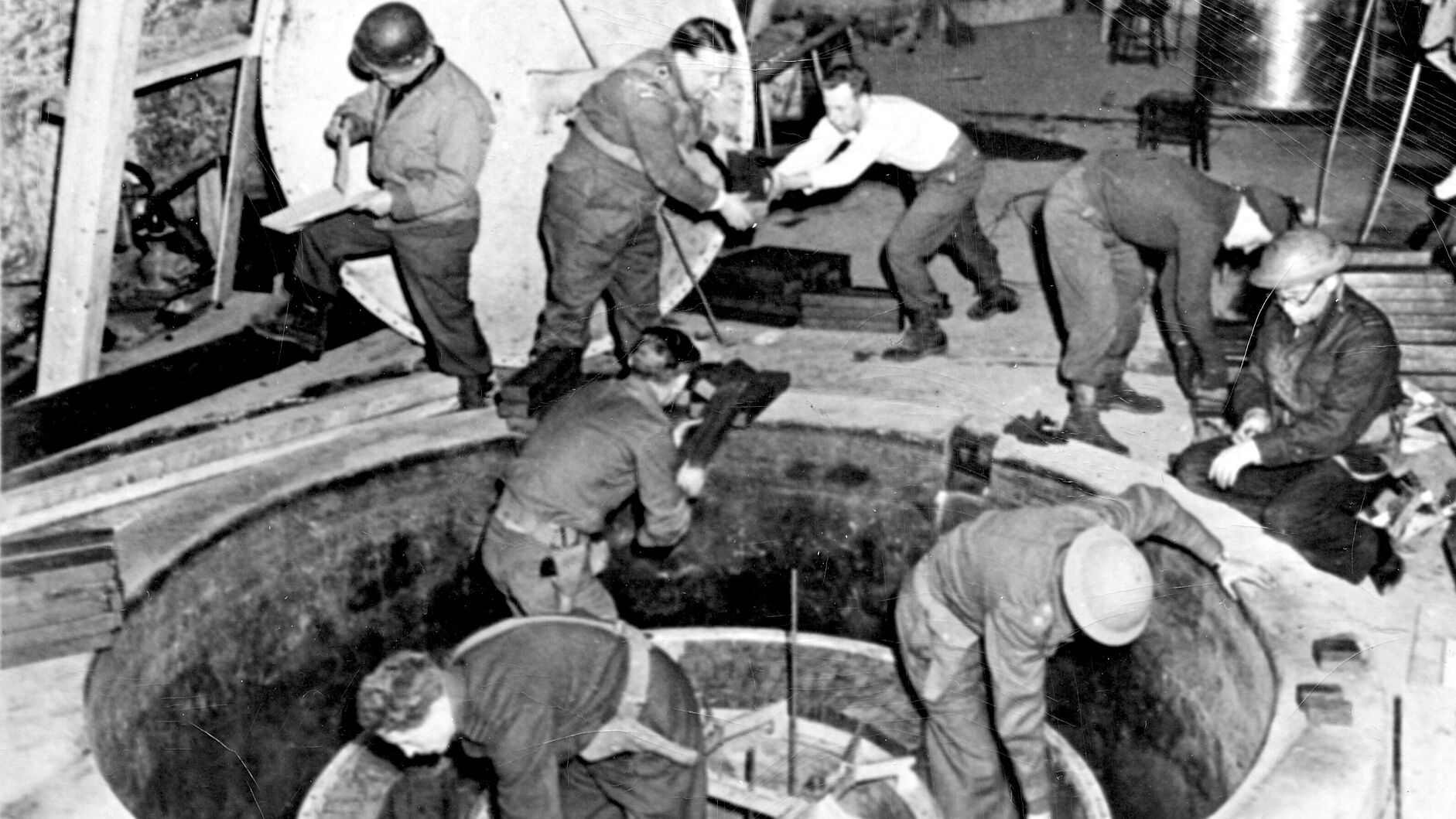 German Uranium Machine 1945 Fotografie. Acht Männer in Uniform, teils mit Helm, verrichten Arbeiten an einem im Boden eingelassen ca. 2 Meter breiten Apparat.