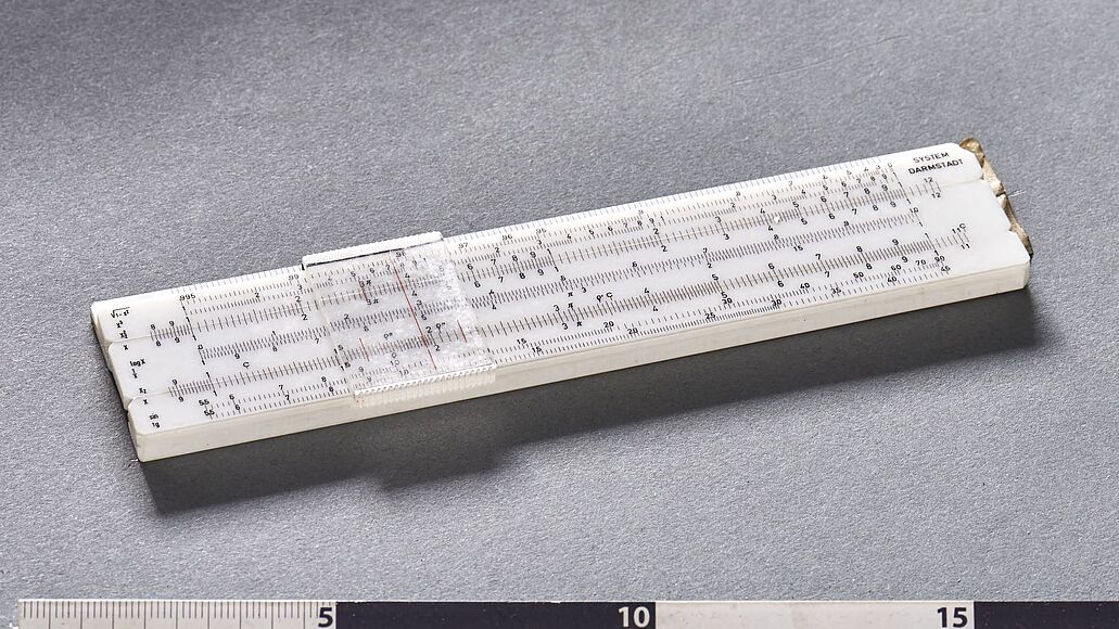 Weißer Quader mit mehreren Skalen vor grauem Hintergrund. Plastikplatte verschiebbar darüber angebracht