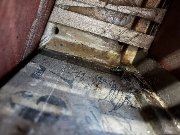 Nahaufnahme des Innenraums: eingerissene Leinen- oder Juteschichten, staubige Unterlage mit Schmier- und Bleistiftkratzern, in warmen Braun- und Rottönen.