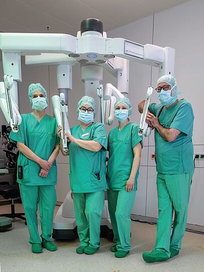Fotografie von vier Personen in OP-Begleidung und OP-Masken vor und unter einem weißen massiven Gestell mit vier Armen (OP-Roboter).