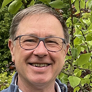 Ein lächelnder Mann mit dunkler Brille, graubrauen Haaren und hellblauem Hemd unter einem blauen Reißverschluss-Pullover, vor grünen, blättrigen Sträuchern im Garten.