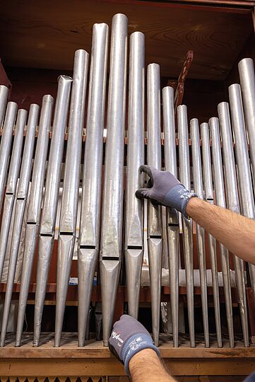 Fotografie von einer Reihe Orgelpfeifen. Zwei Hände in Handschuhen greifen gerade an eine der Pfeifen.