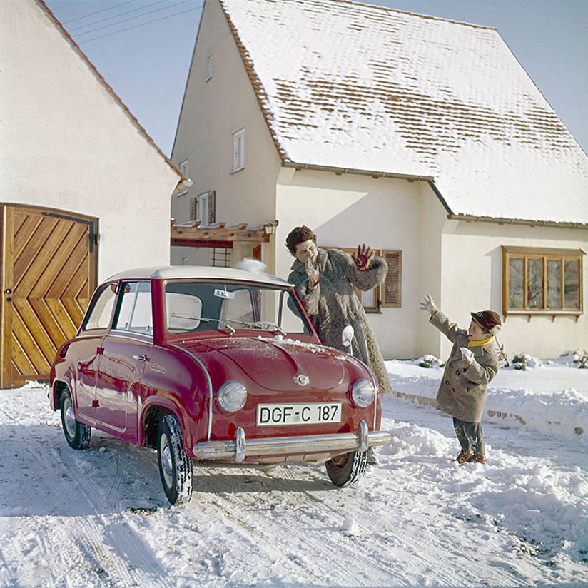 Vor Reihenhaus steht ein rotes Goggomobil in einer Winterlandschaft. Danben stehen Mutter und Kind. Das Kind wirft mit einem Schneeball.