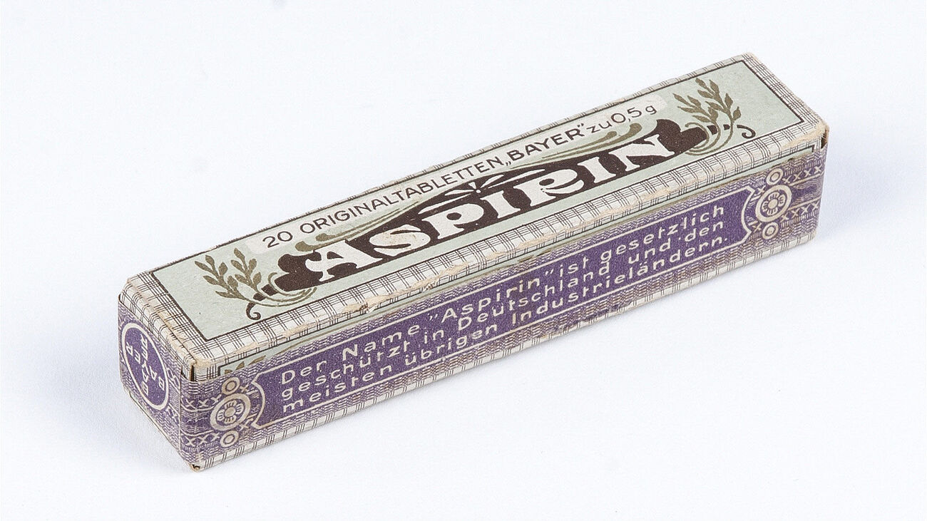 Kleine rechteckige Pappschachtel für „Aspirin“ der Marke Bayer, im Stil des frühen 20. Jahrhunderts. Die Schachtel zeigt dekorative Muster in Lila und Beige, den Schriftzug „Aspirin“ oben sowie Hinweise zu „20 Originaltabletten, Bayer, zu 0,5 g“. Die Seiten tragen zusätzliche Produktangaben in deutscher Sprache.