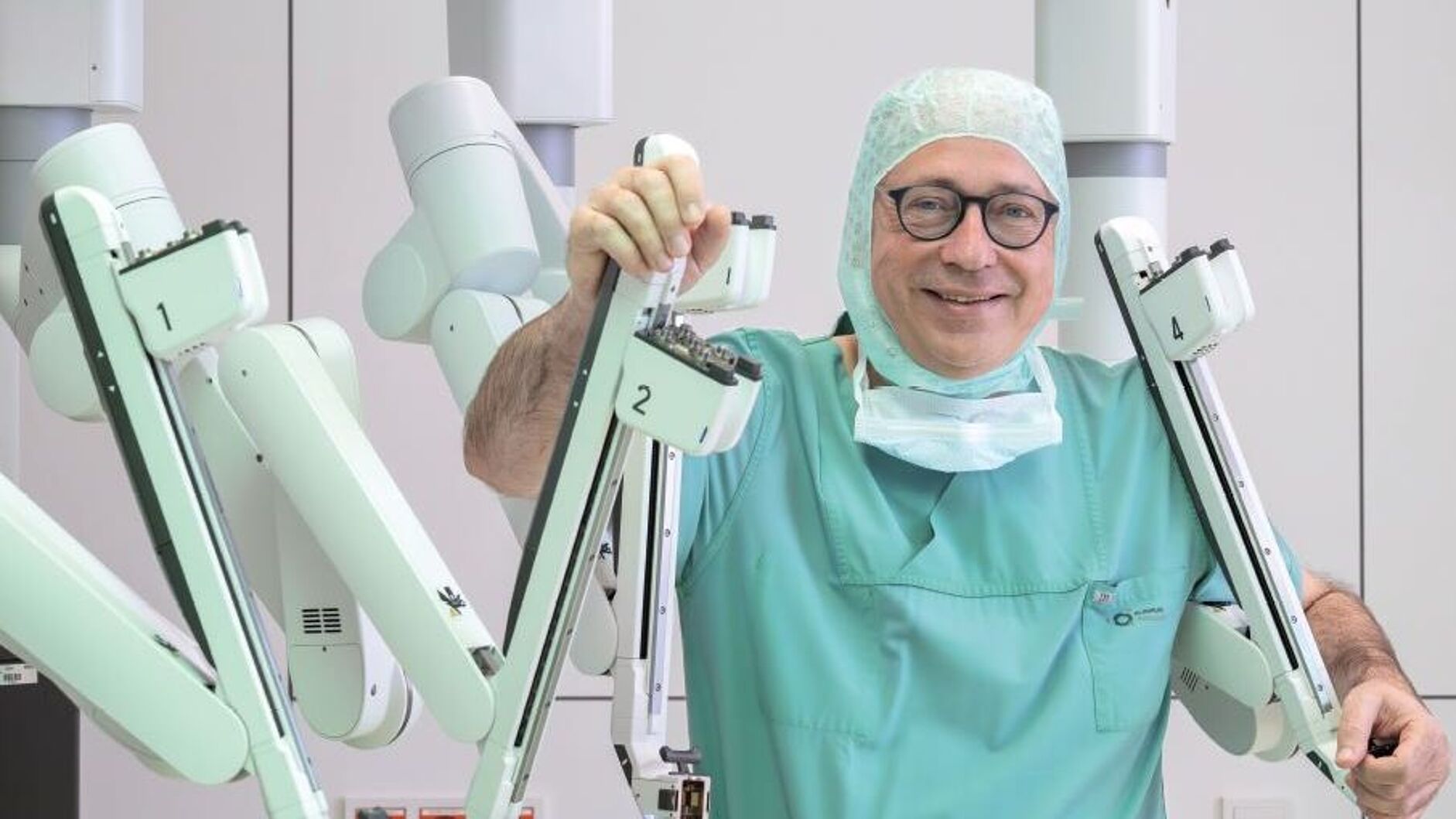 Fotografie von einem Mann mit Brille, abgenommener OP-Maske und OP-Bekleidung. Er hält sich an einem weißen massiven Gestell mit mehreren Armen (OP-Roboter) fest.