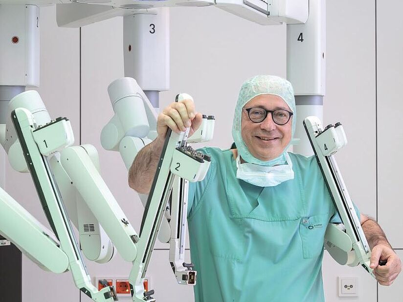 Fotografie von einem Mann mit Brille, abgenommener OP-Maske und OP-Bekleidung. Er hält sich an einem weißen massiven Gestell mit mehreren Armen (OP-Roboter) fest.