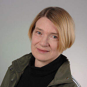 Porträt einer Frau mit kinnlangem blondem Bob, die eine olivgrüne Jacke mit weißen Seitenstreifen über einem schwarzen Rollkragenpullover trägt; Gesicht unscharf/abgedeckt.