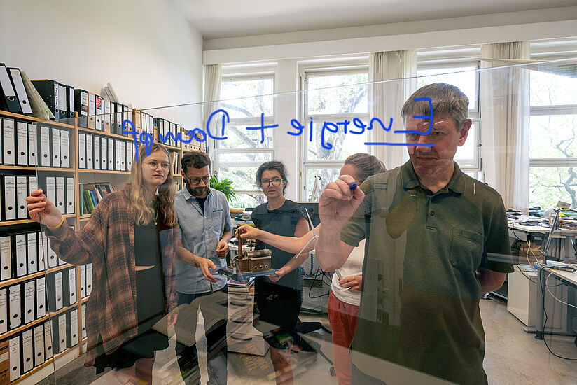 Büroraum. Im Vordergrund schreibt ein Kurator "Energie und Dampf" auf eine Scheibe, im Hintergrund wird sich mit einem technischen Modell beschäftigt.