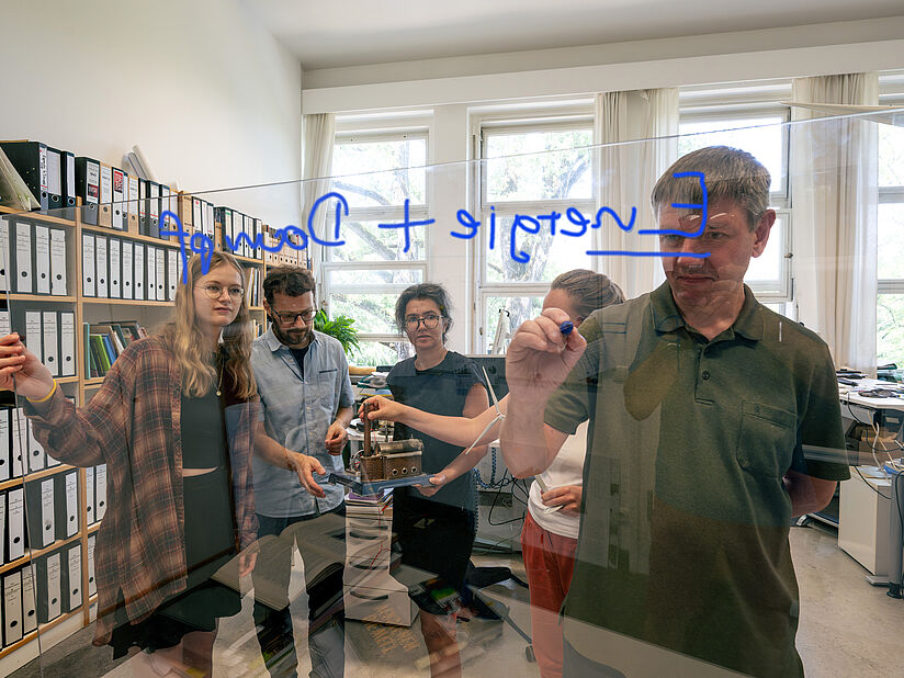 Büroraum. Im Vordergrund schreibt ein Kurator "Energie und Dampf" auf eine Scheibe, im Hintergrund wird sich mit einem technischen Modell beschäftigt.