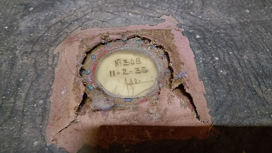 Nahaufnahme eines runden, verblassten Etiketts in einem ausgefransten Stofffenster auf einer dunklen, rissigen Holzfläche; das Etikett zeigt handschriftliche Notizen „N: 348“ und das Datum „11-2-35“ sowie eine Signatur, umgeben von kleinen Nägeln und zerfetztem rosa-braunem Leinen.