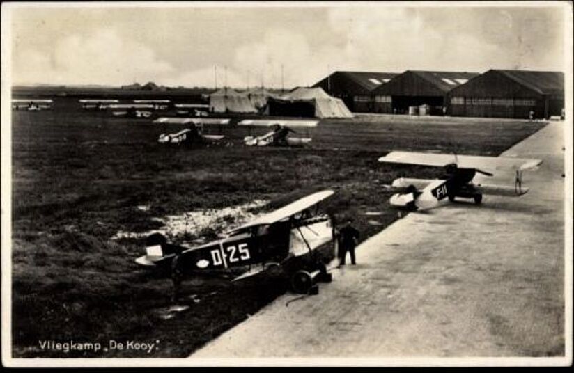 Schwarzweiß-Foto eines historischen Flugplatzes mit mehreren Doppeldeckern und Flugzeugen auf dem Grasfeld und einem betonierten Rollweg; im Vordergrund ein Doppeldecker mit Kennung "D-25" und daneben eine einzelne stehende Person, im Hintergrund Zelte und große Hangars; Beschriftung unten links: "Vliegkamp De Kooy".