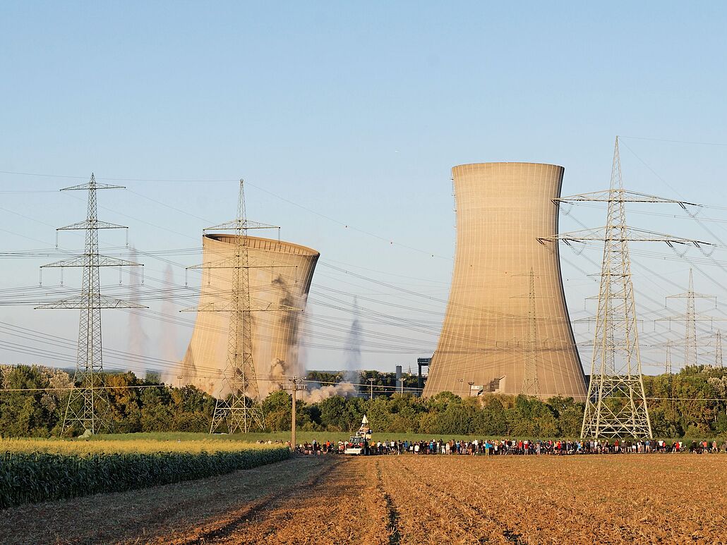 Im Vordergrund sind ein Feld, Strommasten und in einiger Entfernung eine Ansammlung von Menschen. Im Hintergrund: zwei Kühltürme eines Atomkraftwerks, der linke bricht nach rechts ein, am Boden ist Staub von der Sprengung zu sehen.