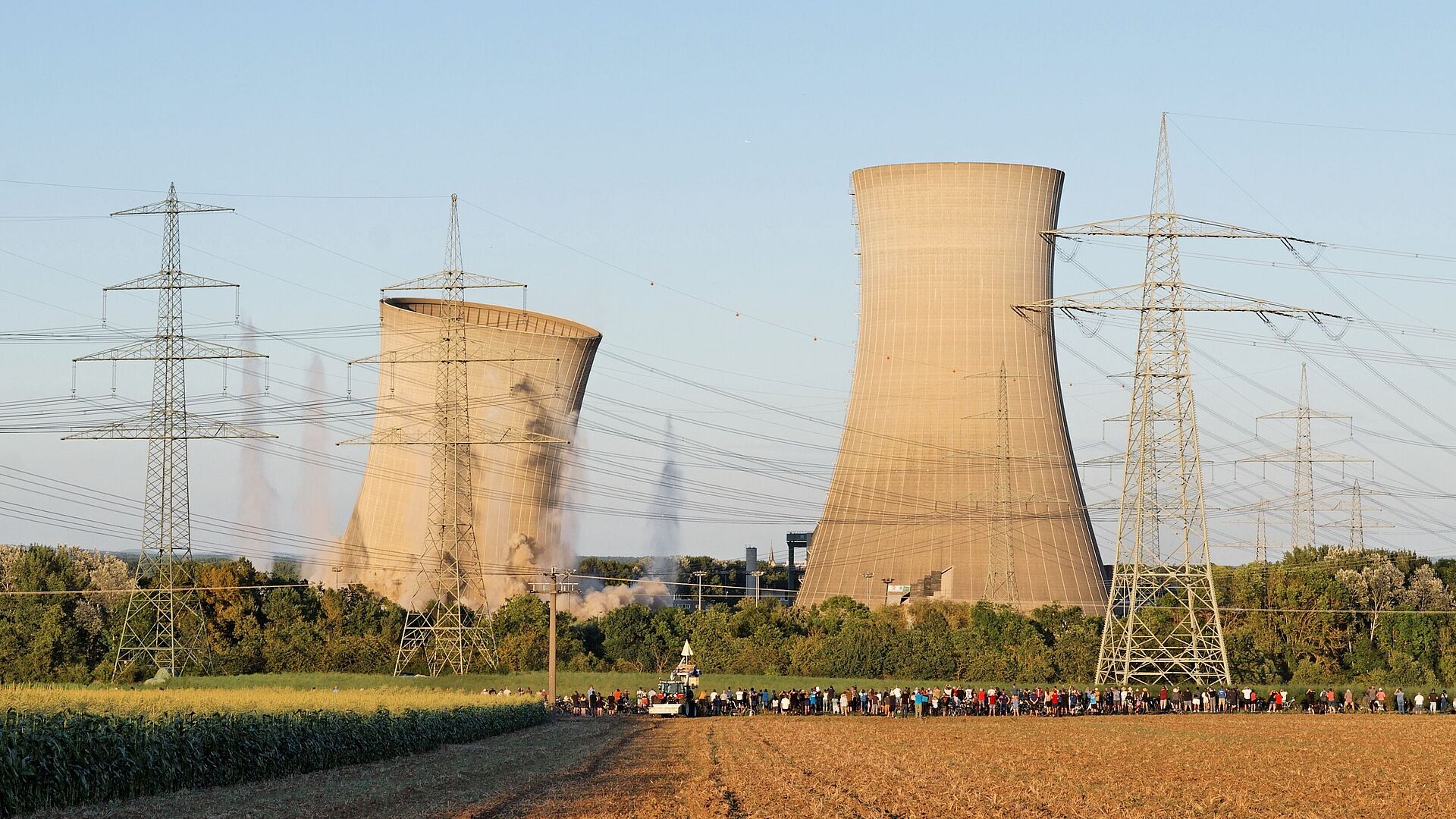 Im Vordergrund sind ein Feld, Strommasten und in einiger Entfernung eine Ansammlung von Menschen. Im Hintergrund: zwei Kühltürme eines Atomkraftwerks, der linke bricht nach rechts ein, am Boden ist Staub von der Sprengung zu sehen.