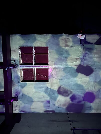UV-Licht beleuchteter Flügel mit vier kleinen Öffnungskästen in einem 2x2-Raster, davor Schatten und violett-blau-grüne Lichtflecken, links sichtbare Metallträger.