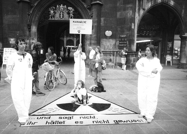 Schwarz-weiß Fotografie. Drei Frauen in weißen Schutzanzügen stehen an den Ecken eines auf dem Boden aufgelegten Kernkraftsymbols. Die hintere hält ein Holzschild "Mahnwache". Schriftzug am Boden: "... und sagt nicht, ihr hättet nichts gewusst." Passanten im Hintergrund.