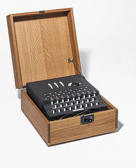 Holzkiste mit offenem Deckel, darin eine schwarze, mechanische Chiffriermaschine/Enigma-ähnliche Schreibwalzen-Einheit mit kreisförmigen Tasten und Metallhebeln, Kabel im vorderen Fach sichtbar.
