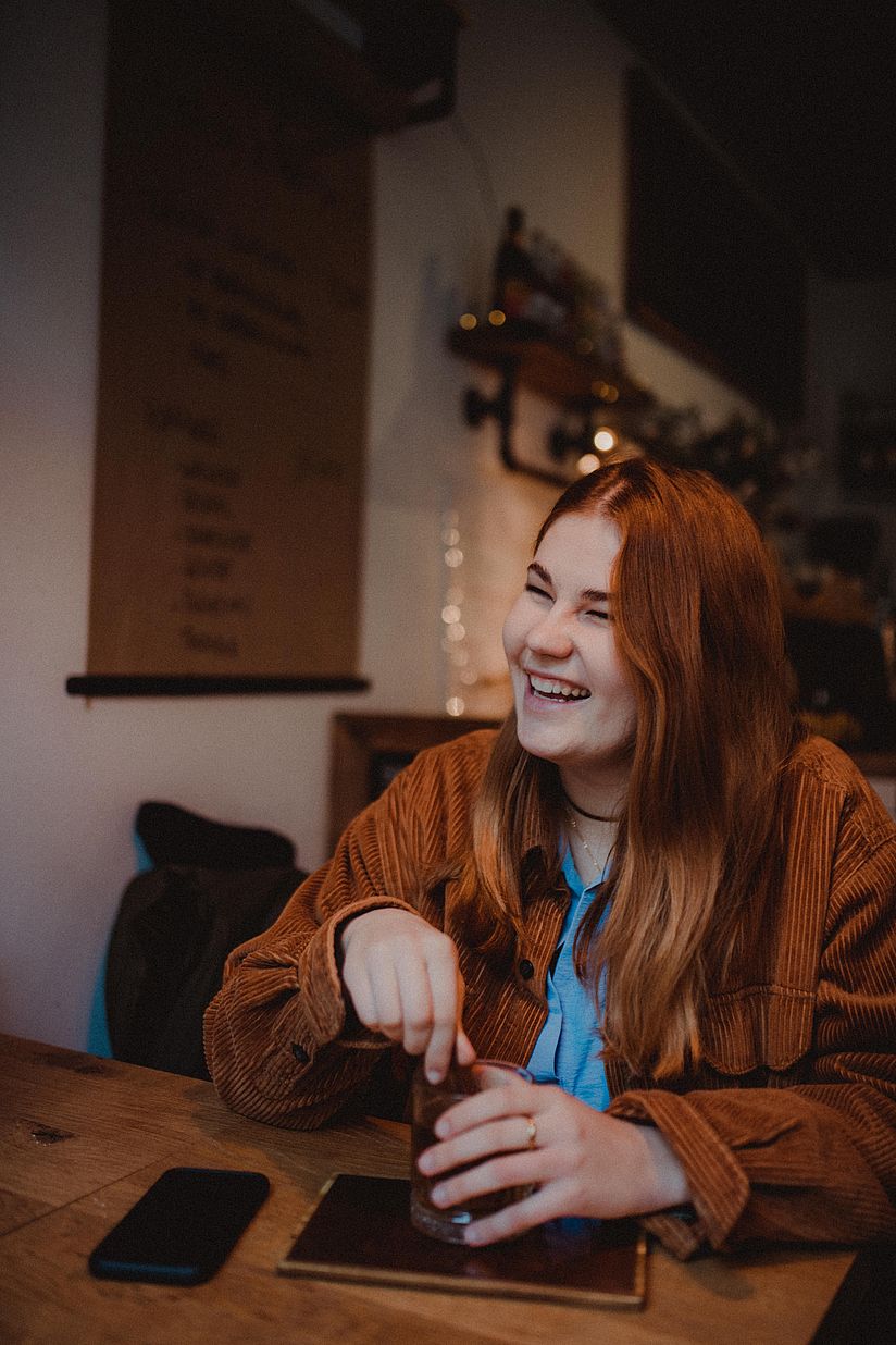 Lisa-Sophie Scheurell hat lange glatte Haare und ein freundliches Lächeln. Sie sitzt an einem Tisch mit einem Getränke, vor sich ein Smartphone und ein Tablet. 