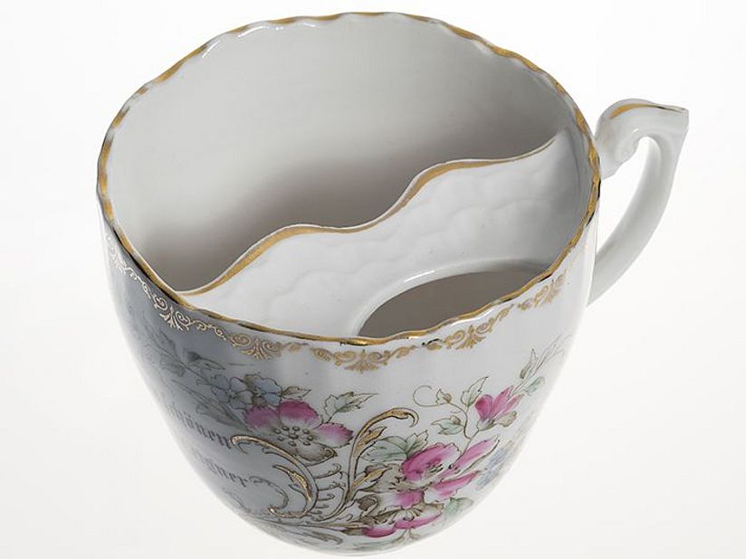 Barttasse mit Goldstaffagen und Spruch „Zu schonen Deinen schönen Bart, nimm diese Tasse eig´ner Art.“. Die Tasse wurde um 1900 von der schlesischen Porzellan-Manufaktur K.P.M. Carl Franz Krister Waldenburg hergestellt.