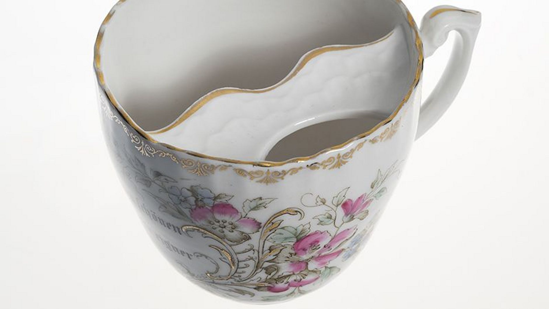 Barttasse mit Goldstaffagen und Spruch „Zu schonen Deinen schönen Bart, nimm diese Tasse eig´ner Art.“. Die Tasse wurde um 1900 von der schlesischen Porzellan-Manufaktur K.P.M. Carl Franz Krister Waldenburg hergestellt.