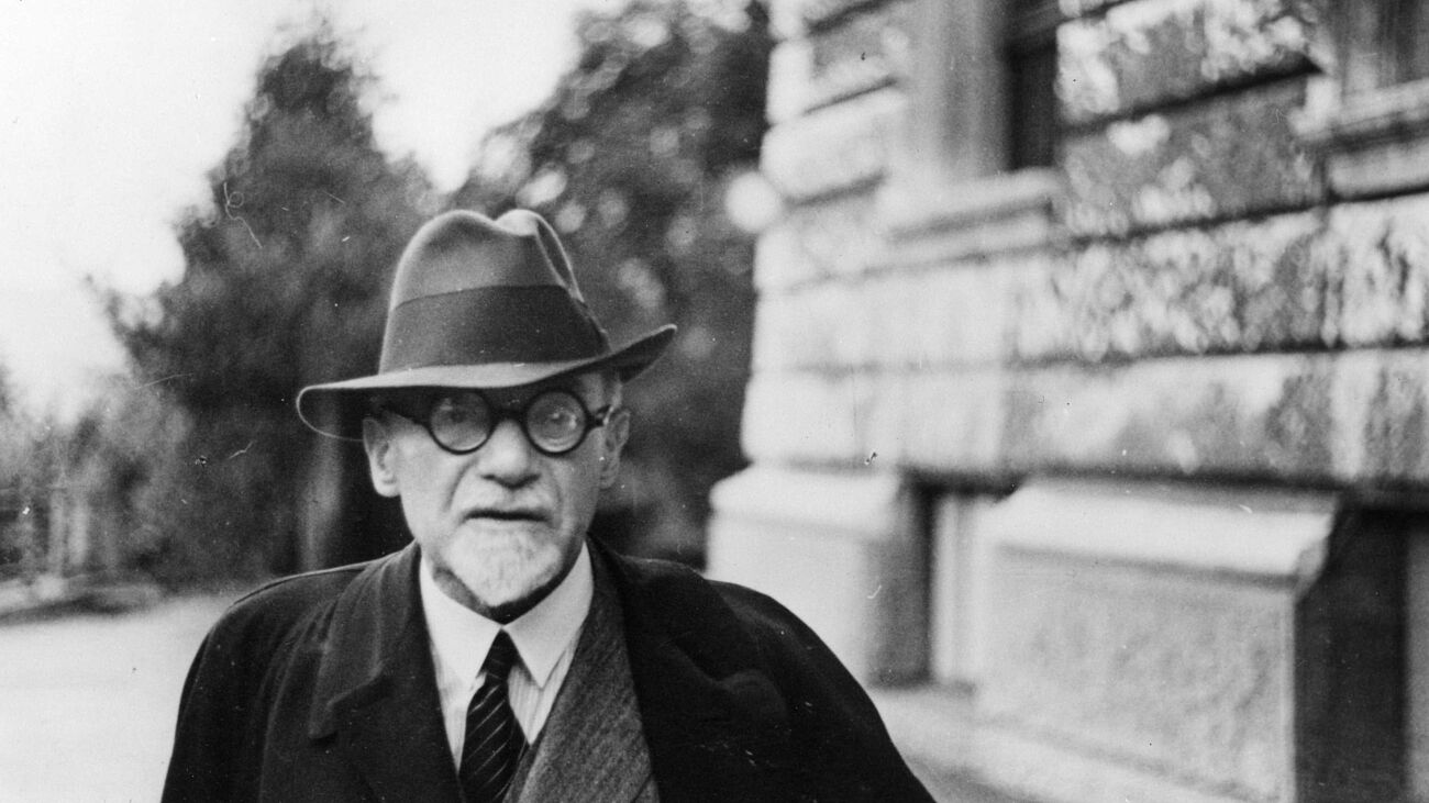 Schwarz-weiß Fotografie. Arnold Berliner mit Hut, Brille und Mantel bis zur Brust sichtbar vor einem verschwommenen Hintergrund (rechts steinernes Gebäude, links und weiter hinten Bäume und Straße).