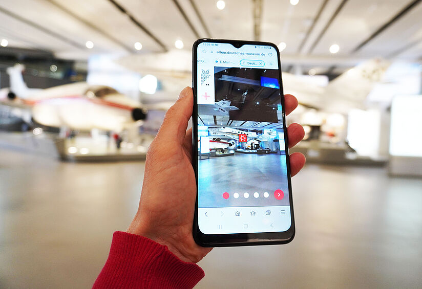 Handy in Hand, der Bildschirm zeigt das gleiche Bild wie vor dem Handy: einen Ausschnitt der modernen Luftfahrt Ausstellung.