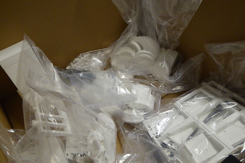 Aufgeklappter Karton voller transparenter Kunststoffbeutel mit vielen weißen Kunststoffteilen und Gehäuseelementen, stapelbare runde und rechteckige Komponenten sichtbar, verstreut verpackt.