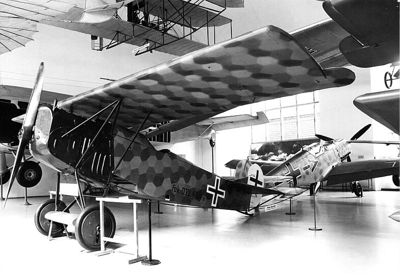 Schwarzweißfoto eines historischen Doppeldecker-Kampfflugzeugs mit sechseckiger Tarnung und deutschem Balkenkreuz im Museum, Seitenansicht im Hangar mit weiteren Flugzeugen im Hintergrund.