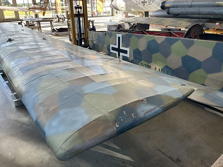 Nahaufnahme eines reparierten Flugzeugflügels in einer Halle: bemalte Stoffbespannung mit polygonalem, mehrfarbigem Tarnmuster in Grün-, Blau- und Braun­tönen, sichtbaren Flicken und Nähten sowie einem schwarzen Balkenkreuz auf dem Rumpfabschnitt im Hintergrund; Werkstattumgebung mit Werkzeugen und weiteren Flugzeugteilen.