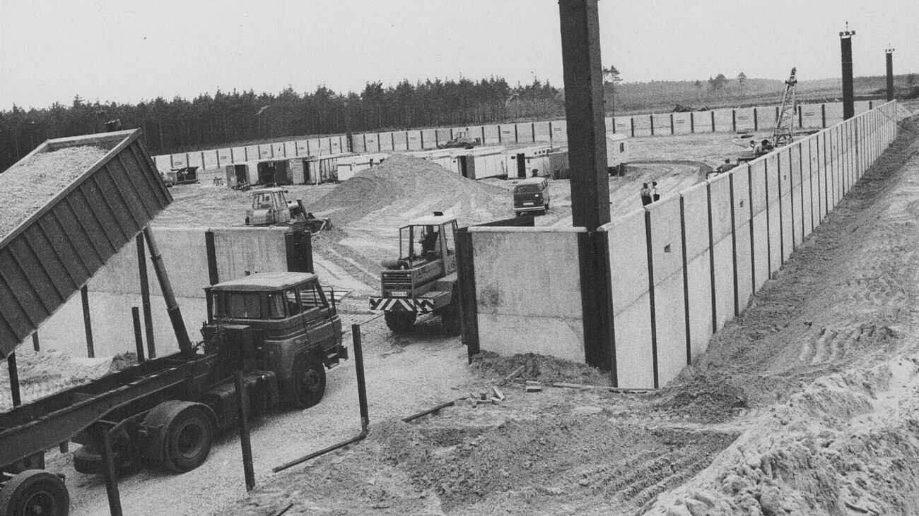 Schwarzweiß Fotografie. Baustelle von Betonmauern umgeben, aufgeschütteter Sand im Inneren und an der Seite. Es fahren Kipplaster, Bagger und ein PKW in die Baustelle. Im Hintergrund Forst.