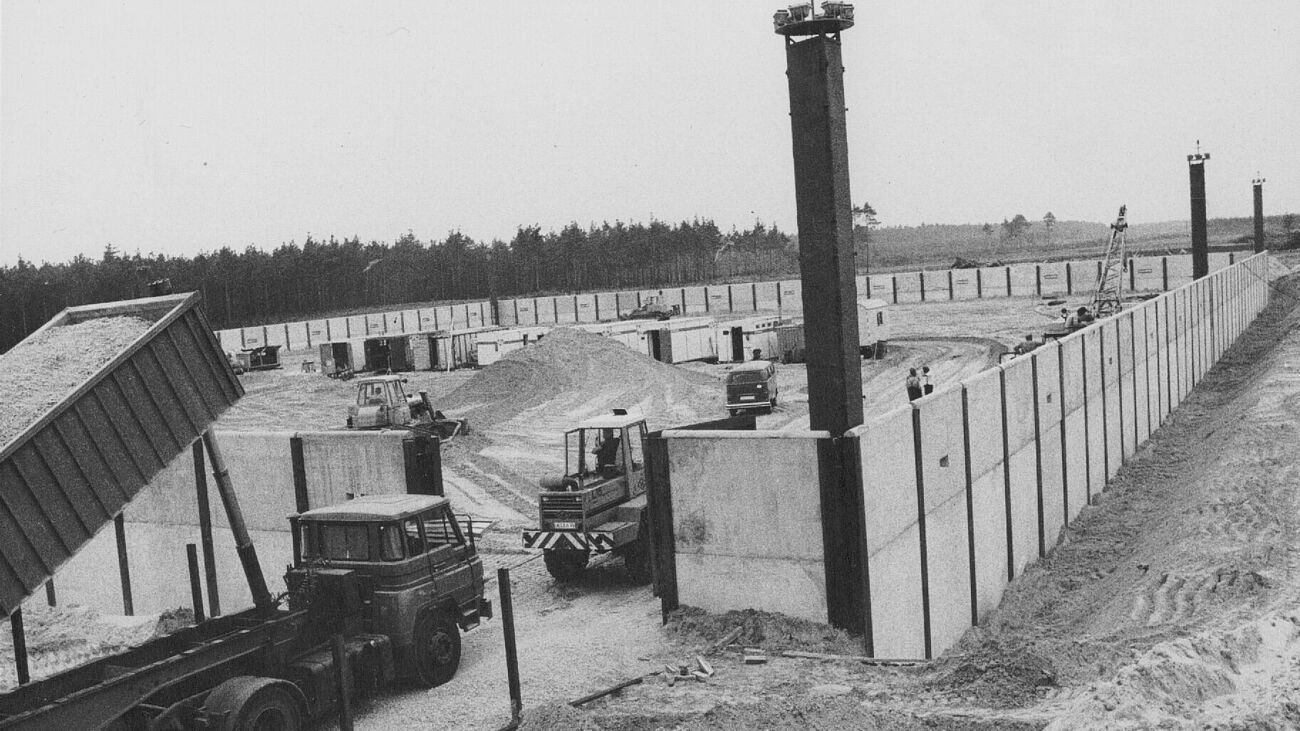Schwarzweiß Fotografie. Baustelle von Betonmauern umgeben, aufgeschütteter Sand im Inneren und an der Seite. Es fahren Kipplaster, Bagger und ein PKW in die Baustelle. Im Hintergrund Forst.