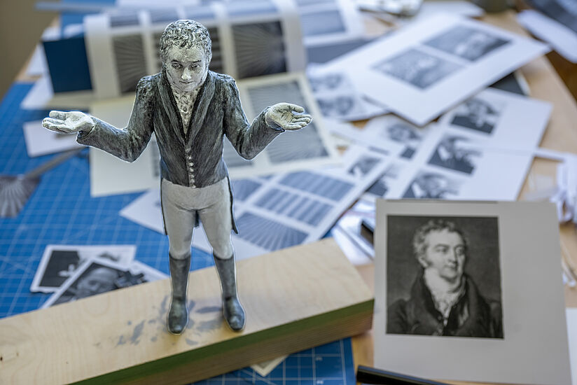 Der in Gehrock und Stiefeln gekleidete Thomas Young als Figur auf einem Tisch, daneben ein Foto des Wissenschaftlers.