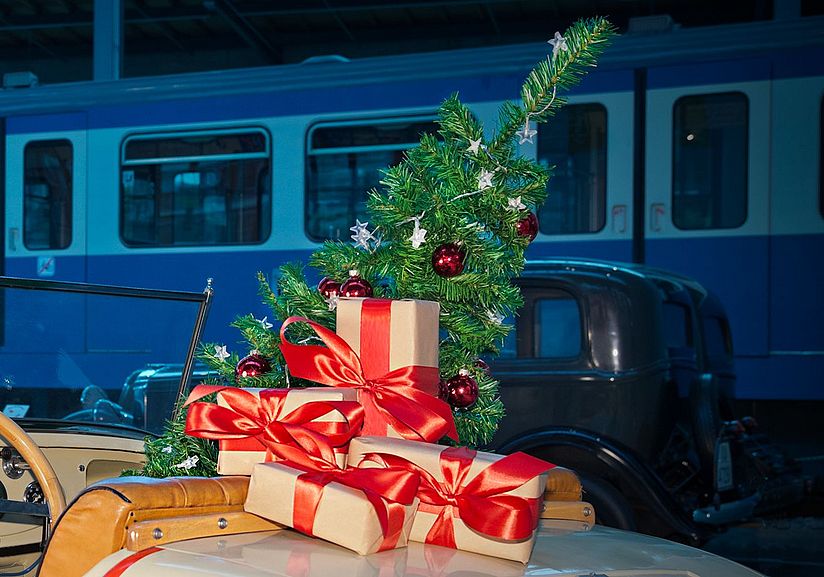Auf einem Oldtimer befinden sich vier verpackte Geschenke mit roter Satinschleife und ein kleiner, geschmückter Weihnachtsbaum.