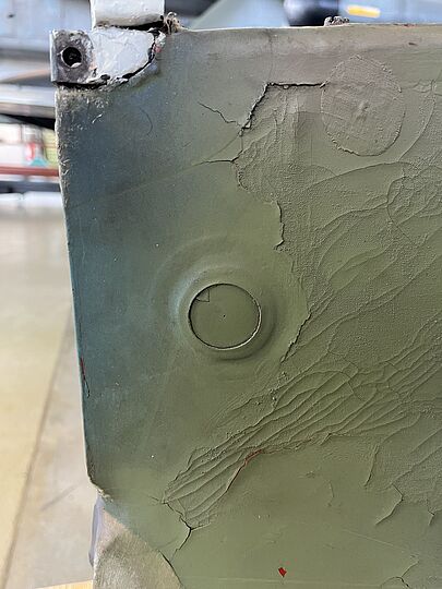 Nahaufnahme einer olivgrün lackierten Metallfläche mit abgeplatzter, rissiger Farbe und strukturierten Spachtelstellen; rundes Blindblech/Abdeckkappe mittig links, Kratzspuren und kleine rote Farbreste, obere Ecke mit beschädigter weiß lackierter Ecke und sichtbarem Schraubenloch.
