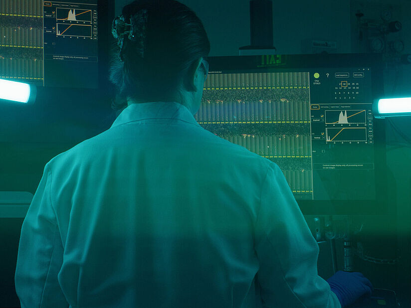 Eine Wissenschaftlerin im Laborkittel betrachtet Daten auf einem Bildschirm in einem blaugrün beleuchteten Labor.